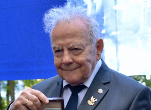 Zdjęcie przedstawia starszego mężczyznę prof. Andrzeja Januszajtisa trzymającego w dłoni dokument oraz medal. W tle fragment ekranu i zieleń.  