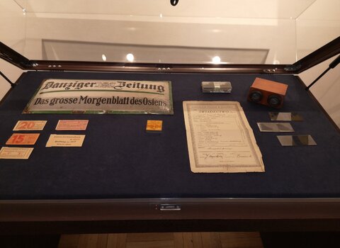 Zdjęcie przedstawia leżącą w gablocie metalową, prostokątną tablicę z napisem Danziger Zeitung i inne mniejsze pamiątki w tym dokument i stereoskop - przedmiot o kształcie aparatu. 