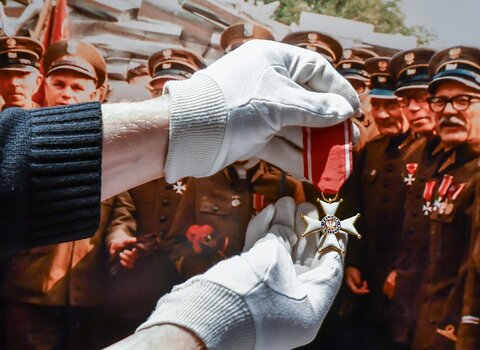 Zdjęcie przedstawia dłonie w rękawiczkach trzymające Order Polonia Restituta. W tle zdjęcie z uroczystości. Widnieją na nim starsi mężczyźni ubrani w mundury. Na piersiach mają odznaczenia. Order trzymany w dłoniach jest podobny do noszonego na mundurze jednego z członków uroczystości, Alfonsa Pillatha (po prawej od dłoni).   