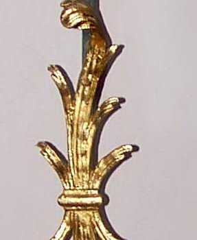 Pionowe zdjęcie przedstawia oryginalny dzwonek. Podłużny pręt z motywami roślinnymi na dwóch końcach. Motywy są złocone.  