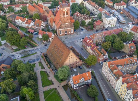 Zdjęcie przedstawia widok na ulice Starego Przedmieścia z lotu ptaka. W centrum znajduje się bryła Wielkiego Młyna ze spadzistym dachem i opływający go Kanał Raduni. Nieco wyżej ulica Wielkie Młyny oraz kościół św. Katarzyny wraz z wieżą kościoła. Poniżej zielone drzewa i Skwer Heweliusza.  