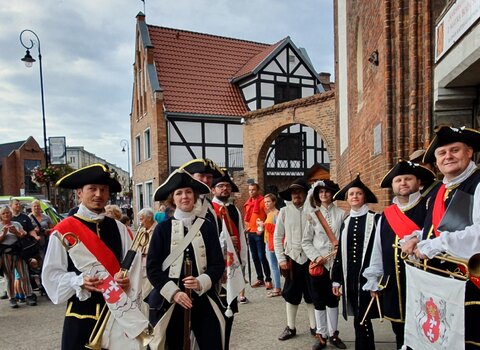 Zdjęcie przedstawia stojące w dwóch rzędach grupkę ludzi przebranych w historyczne stroje. W tle fragment fasady kościoła św. Katarzyny oraz Małego Młyna.  