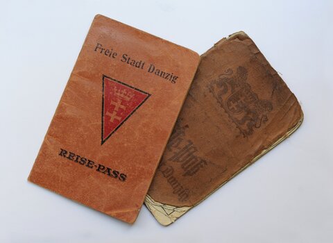 Zdjęcie przedstawia dwa paszporty z okresu Wolnego Miasta Gdańska. Na prostokątnej oprawie trójkąt, a w nim herb Gdańska. Nad nim napis Freie Stadt Danzig. Pod trójkątem napis Reise-pass. 