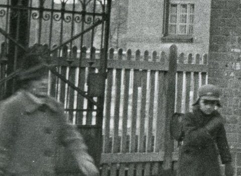 Zdjęcie przedstawia mur a w nim otwartą bramę i w głębi sylwetkę Domu Polskiego przy Wallgasse 16. Przed bramą dwóch przechodniów. Bliżej bramy dziecko w czapce i z plecakiem patrzy w kierunku idącej starszej kobiety w kapeluszu na głowie. Na bramie zawieszone 2 tablice z godłem NSDAP oraz druga z napisem Die Danziger Arbeitsfront.   