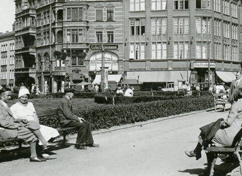 Zdjęcie przedstawia widok na Targ Drzewny. Mężczyźni i kobiety siedzą na ławkach, na prawo od nich obelisk. W centrum zdjęcia fasada budynku.  