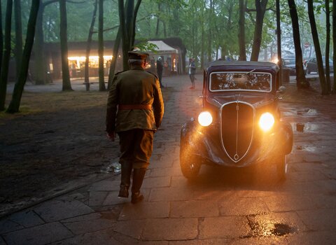 Zdjęcie przedstawia sylwetkę żołnierza Wojska Polskiego z czasów II wojny światowej. W jego kierunku jedzie zabytkowe auto z zapalonymi reflektorami.  