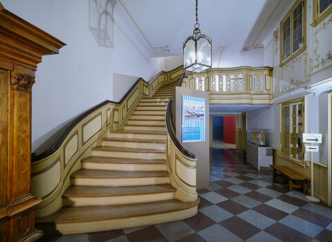 Zdjęcie przedstawia klatkę schodową wewnątrz Domu Uphagena. Na lewo fragment mebla, w centrum schody. Po prawej stronie ściana z okiennicami i wejście do kasy. Z sufitu zwisa stara lampa.   