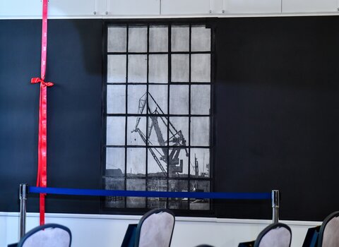 Zdjęcie przedstawia grafikę przesłoniętą wstęgą. W centrum imitacja starego prostokątnego okna podzielonego na mniejsze kwadratowe fragmenty. W oknie sylwetka portowego żurawia. 