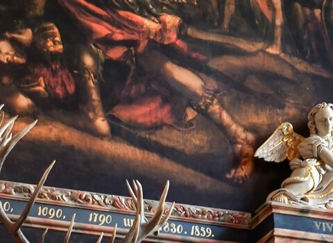 Zdjęcie przedstawia replikę leżącego jelenia nad Ławą św. Rajnolda w Wielkiej Hali Dworu Artusa. Wokół niej liczne zdobienia i kolumny. Nad jeleniem fragment obrazu.  