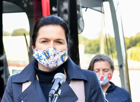 Na zdjęciu prezydent Gdańska Aleksandra Dulkiewicz przemawia przed mikrofonem w maseczce. W tle sylwetka tramwaju.  