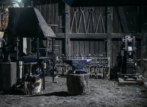 Zdjęcie przedstawia wnętrze Kuźni Wodnej w Oliwie. Na pierwszym planie po lewej okap paleniska, metalowe kowadło i przyrząd. Za nimi zawieszone na drewnianej konstrukcji metalowe narzędzia.  