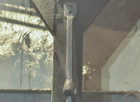Zdjęcie przedstawia zawieszone na słupie serce dzwonu. Jest to metalowy, podłużny przedmiot w kształcie pręta. Na ok 1/3 długości od dołu ma wyraźne zgrubienie w kształcie kuli. Na samej górze metalowy, płaski uchwyt z dziurą. 