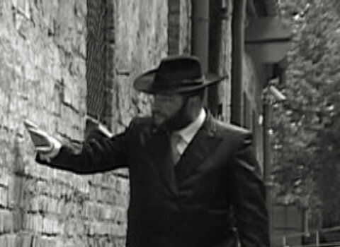 Na zdjęciu w odcieniach szarości spacerujący mężczyzna dotykający ręką ceglanego muru.