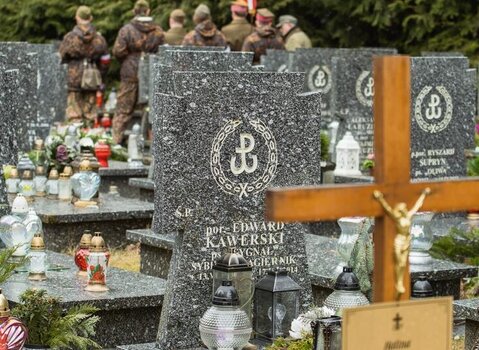 Opis: Zdjęcie kolorowe. W kadrze na pierwszym planie po prawej drewniany krzyż, za nim kamienne nagrobki. W centrum nagrobek porucznika Edwarda Kawerskiego. Nad napisem symbol Polski Walczącej otoczony laurem. W tle rozmyte sylwetki harcerzy. 