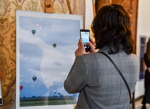Zdjęcie. Kobieta stojąca plecami do widza fotografuje telefonem komórkowym fotografię przedstawiającą wzlatujące w zachmurzone niebo balony. 