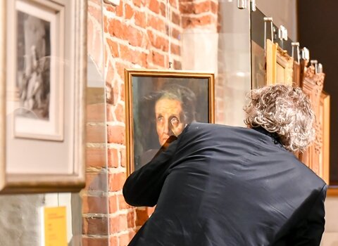 Zdjęcie. Zwiedzający wernisaż wystawy we wnętrzach Galerii Palowej. Trzech mężczyzn ogląda obraz.