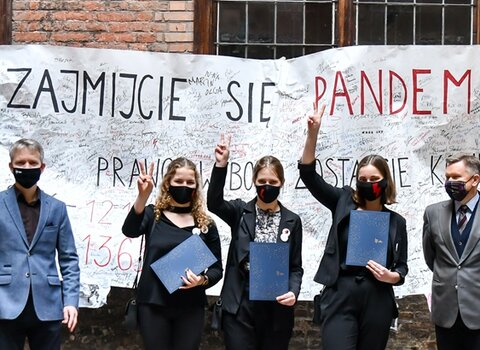 Zdjęcie. Dziedziniec. Od Janusz Marszalec, lewej Julia Landowska, Aleksandra Łoboda i Marianna Łoboda oraz Waldemar Ossowski. Za nimi transparent z napisem "Zajmijcie się pandemią". Kobiety trzymają uniesione dłonie z symbolem zwycięstwa.  