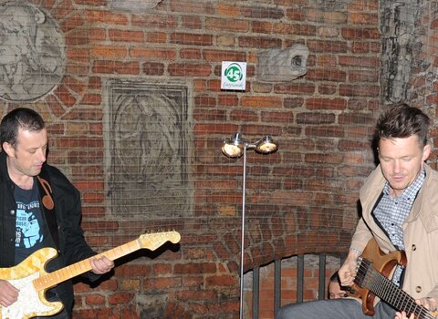 Koncert na dziedzińcu Muzeum Bursztynu. Po lewej Tymon Tymański, gra na gitarze z żółtego bursztynu. Po prawej Wojciech Mazolewski, gra na zwyczajnej gitarze. Obok leżą kable, wzmacniacz, futerał.
