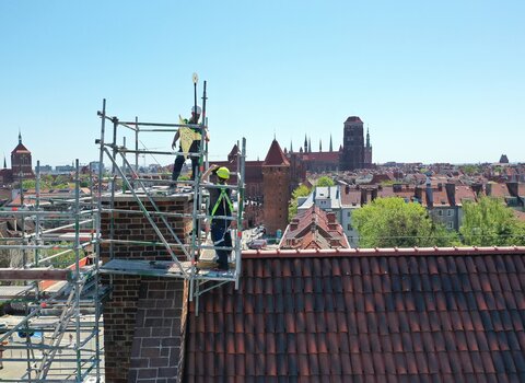 Dwaj mężczyźni stoją na rusztowaniu wokół komina Wielkiego Młyna. Montują złotą chorągiew wiatrową o kształcie topora.