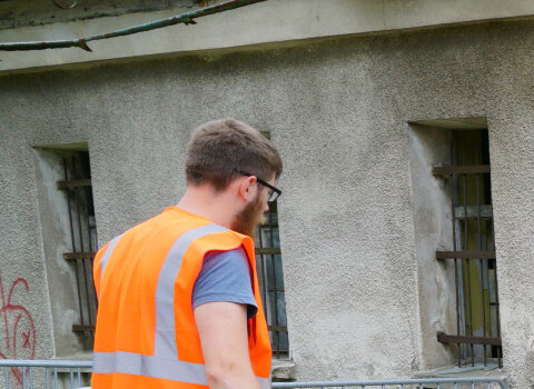 Zdjęcie. Przestrzeń. Budynek z niewielkimi zakratowanymi oknami, pokryty szarym tynkiem. Poniżej wykop. Archeolog patrzy w wykop i odsłoniony fragment fundamentów koszar napoleońskich. 