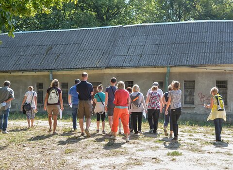 Zdjęcie. Przestrzeń. Ludzie przed budynkiem koszar napoleońskich. Patrzą na dach pokryty eternitem. W tle drzewa.