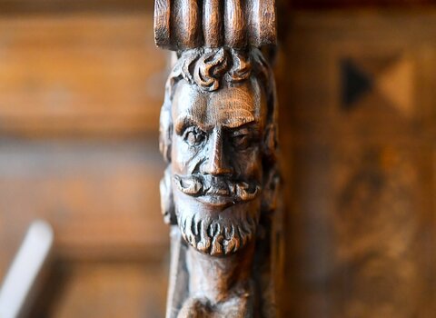 Zdjęcie. Fragment ławy. Bok (uszko) dekorowany rzeźbą przedstawiają brodatego mężczyznę. 