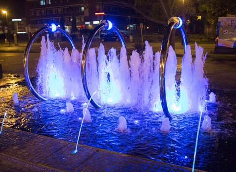 Zdjęcie. Wieczór. Fontanna Heweliusza - iluminowany pokaz wodny. W tle ulica.  