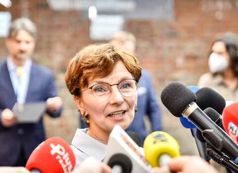 Zdjęcie. Renata Adamowicz. Wokół mikrofony. W tle ceglany mur i ludzie. 