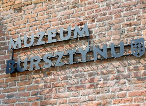 Zdjęcie. Szyld z napisem Muzeum Bursztynu. Obok herb Gdańska. W tle ceglana elewacji Wielkiego Młyna. 