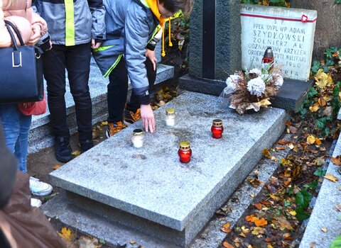 Zdjęcie. Uporządkowany nagrobek ze zniczami. W kadrze - lewym rogu ludzie. 