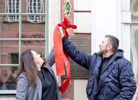 Zdjęcie. Anna Kriegseisen (z lewej) oraz Bartosz Nawacki (z prawej) rozwiązują wstęgę wokół uchwytu repliki dzwonka. W tle fasada Domu Uphagena.