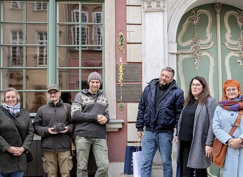 Zdjęcie. Przed fasadą Domu Uphagena. Od lewej Ewa Szymańska, Remigiusz Pacer z łupą żelaza, Grzegorz Ciechomski, Bartosz Nawacki, Anna Kriegseisen oraz Katarzyna Darecka.   