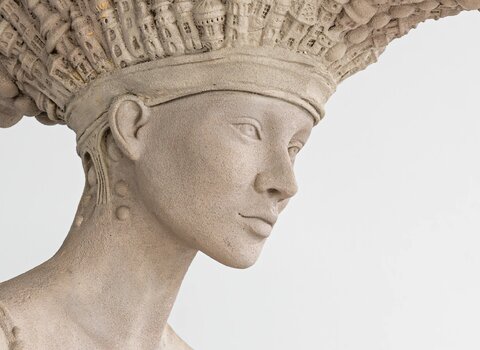 Wizerunek dzieła sztuki. Rzeźba "Architektoniczna" Tomasza Sętowskiego. Postać kobiety z kapeluszem składającym się z budynków.   