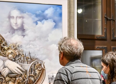 Zdjęcie. Mężczyzna i kobieta patrzą na obraz. Z chmur otaczających bajkowe miasto wyłania się postać Mony Lisy. 