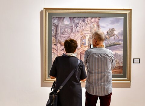 Zdjęcie. Kobieta i mężczyzna patrzą na obraz. Widoczny fragment budynku i postać ujeżdżająca konia.
