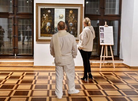 Obraz przedstawiający 2 osoby. Mężczyzna ubrany cały na biało i kobieta stoją na sali sali wystawowej. Patrzą na obraz - tryptyk - przedstawiający wnętrze księgarni.