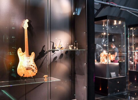 Zdjęcie. Fragment wystawy. Po lewej wisi gitara elektryczna wykonana z bursztynu bałtyckiego. Po prawej gabloty z bursztynowymi przedmiotami w tle.