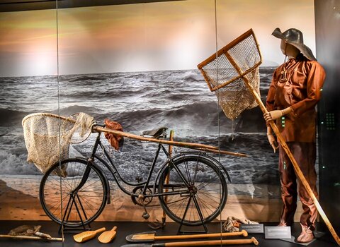 Zdjęcie. Fragment wystawy stałej. Poławiacz bursztynu z kaszorkiem (przyrządem zakończonym siatką). Obok stary rower. W tle Morze Bałtyckie.  