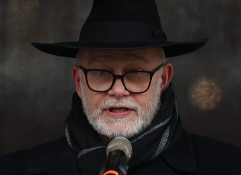 Mężczyzna ubrany na czarno w czarnym kapeluszu przemawia do mikrofonu. Na lewym ramieniu ma materiałową opaskę, w prawej ręce trzyma kartki. To Michał Samet, przewodniczący Gminy Wyznaniowej Żydowskiej w Gdańsku. 