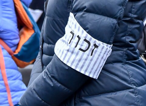 Zbliżenie na rękaw kurtki. Na ramieniu znajduje się materiałowa opaska w biało-szare pasy i napis po hebrajsku.
