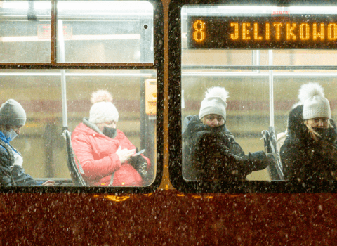 Zdjęcie. Fragment tramwaju z dwoma oknami. Wewnątrz siedzą 4 osoby z maseczkami zakrywającymi częściowo twarz. Patrzą  w telefon, albo w szybę (w kierunki widza). W prawym oknie u góry napis "8 Jelitkowo". Pada śnieg.   