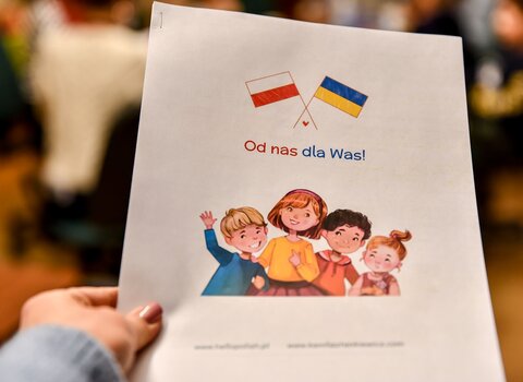 Zdjęcie. Dziecięce dłonie trzymają kartkę z flagami Polski i Ukrainy. Na kartce napis "Od nas dla Was". 