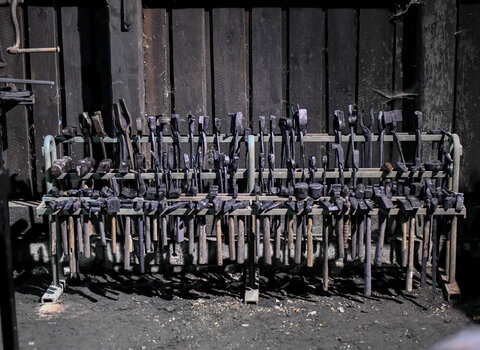 Zdjęcie przedstawia narzędzia kowalskie, szczypce, młoty i inne, umieszczone na czterech poręczach metalowej ramy. W tle drewniane, ułożone poziomo deski - jedna ze ścian Kuźni Wodnej w Oliwie. 