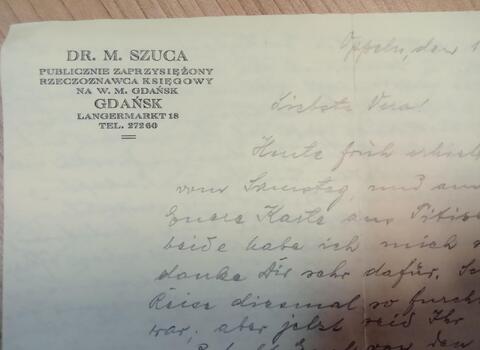 Zdjęcie. Fragment listu na papierze firmowym. W lewym górnym rogu dane adresowe Michała Szucy w języku polskim. Pismo odręczne niżej. Treść w języku niemieckim. 