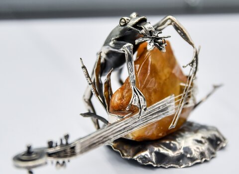 Na zdjęciu srebrna figurka żaby grającej na poziomo ułożonym kontrabasie. Korpus instrumentu wykonany jest z ciemno-miodowej bryłki bursztynu. 