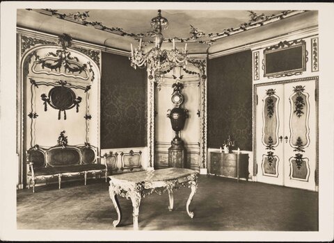 Fotografia czarno-biała w formie pocztówki ukazuje wnętrze domu Uphagena - widok na południowo-wschodnią część salonu. W tle za stołem, zdobiony zegar. Po jego lewej sofa. Po prawej krzesło, a obok niego dekorowane drzwi. 
