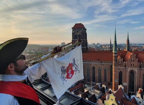 Zdjęcie przedstawia trębacza ubranego w historyczny strój. Do trąbki przymocowany chwytak, na nim flaga a na niej z lwy trzymające herb Gdańska. W tle zabudowania oraz Bazylika Mariacka.  