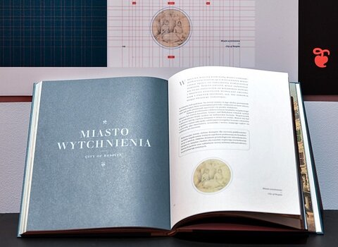 Wnętrze katalogu miasto romantyków. Nad książką plansza pokazująca siatkę z liniami wprowadzonymi przez projektanta.  
