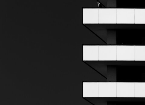 Zdjęcie czarno białe. Nowoczesny budynek. Prawa część zdjęcia jest cała czarna, po prawej widać białe poziome pasy, płyty osłaniające balkony. Na jednym z nich widać sylwetkę człowieka, słabo wyraźną.