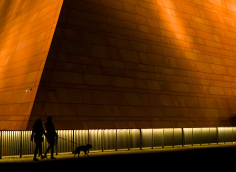 Zdjęcie. Wieczór, noc. Dwie osoby z psem idą nabrzeżem Motławy. Idą na tle budynku, siedziby Muzeum Drugiej Wojny Światowej. Widać naroże budynku o kształcie piramidy. Jest jasno oświetlony od dołu reflektorami.